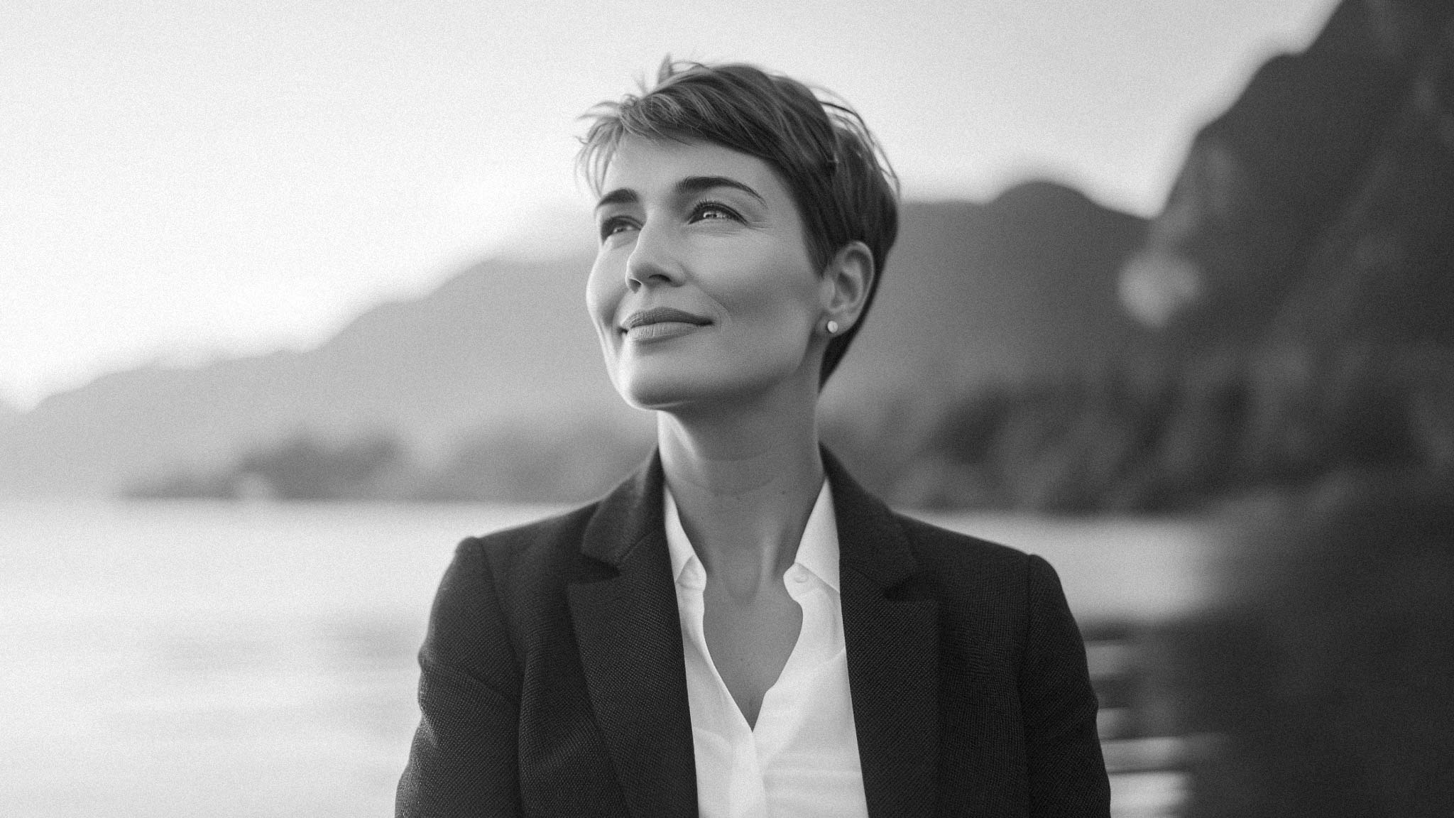 A vaugely AI-looking 40ish woman stares up and to the left with a mountain and ocean blurred backdrop behind her